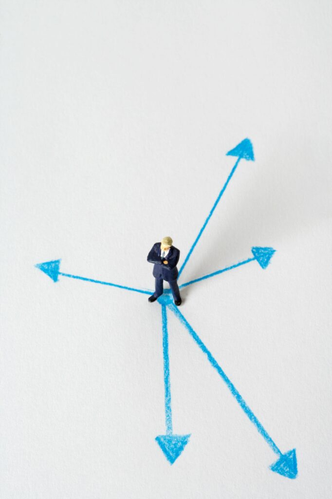 Miniature businessman stands at crossroads of blue arrows on white background, symbolizing choices.