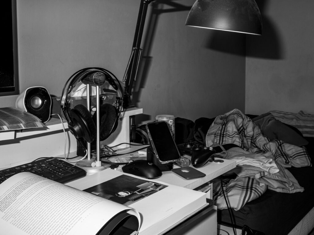 Black and white photo of a cluttered bedroom desk with tech gadgets and reading materials.