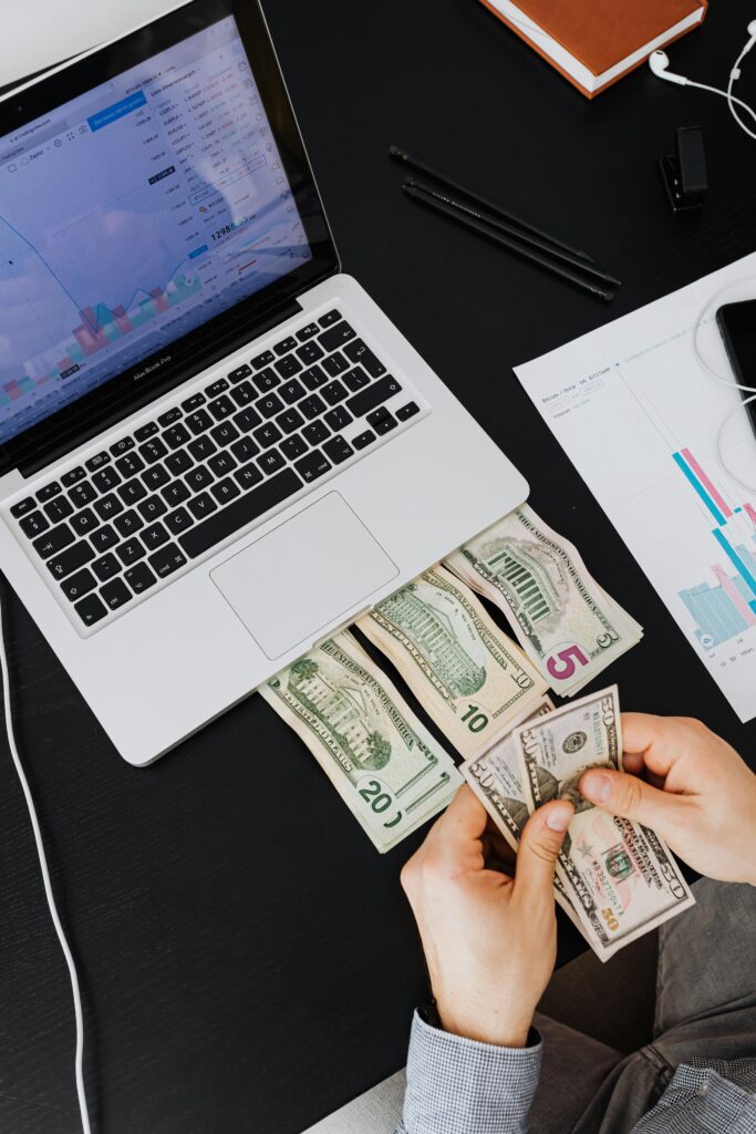 Person counting cash with laptop on desk, financial graphs visible.